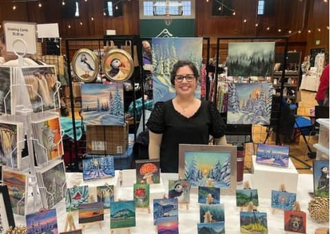 A local artist smiling in her booth of paintings and cards at an indoor holiday market