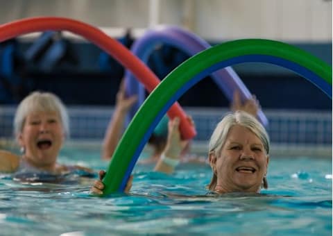 Two smiling women with pool noodles in a water fitness class