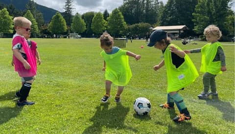 Young children in pinnies chasing a soccer ball on a grass field